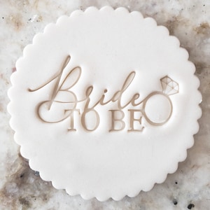 Könnte beinhalten: Runder weißer Ausstecher mit gewelltem Rand. Der Ausstecher ist mit den Worten "Bride to Be" und einem stilisierten Diamantring geprägt.