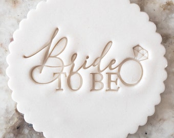 May include: White round cookie cutter with a scalloped edge. The cutter is embossed with the words "Bride to Be" and a stylized diamond ring.