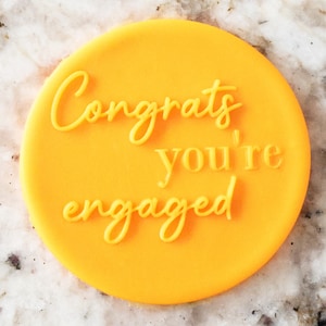 Puede incluir: Un sello redondo de silicona amarillo brillante con el texto "Congrats you're engaged" en una fuente amarilla de escritura.
