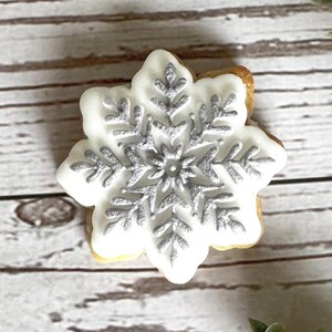 Puede incluir: Una galleta de copo de nieve blanca con glaseado plateado. La galleta está decorada con un diseño de copo de nieve.