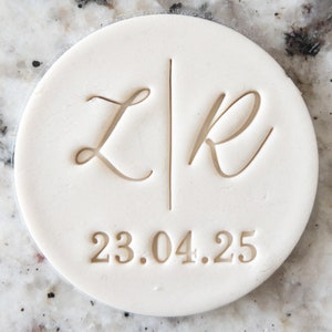 Könnte beinhalten: Ein cremefarbenes, rundes Objekt mit den eingravierten Initialen "L" und "R", getrennt durch einen vertikalen Strich. Unter den Initialen steht das Datum "23.04.25". Das Objekt liegt auf einer hellen, gesprenkelten Oberfläche.