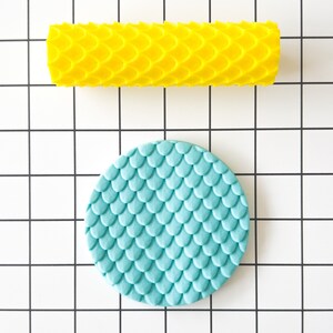 May include: A yellow rolling pin and a light blue round fondant mould, both with a scale pattern. The rolling pin and mould are on a white grid background.