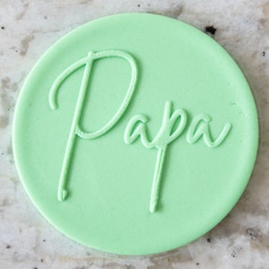 May include: A light green round cookie with the word "Papa" written in white cursive script.