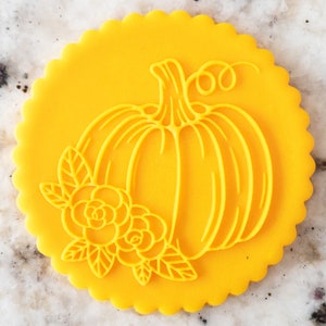 May include: A bright yellow, circular cookie cutter with a scalloped edge. The cutter features a pumpkin design with floral accents. The pumpkin has a stem and a curling vine. The flowers and leaves are also visible.
