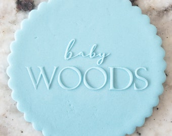 Könnte beinhalten: Ein runder, hellblauer Keks mit gewellten Rändern, verziert mit dem Text "baby WOODS" in weißer Glasur.