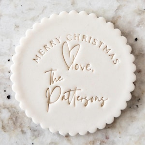May include: White round cookie with a scalloped edge, decorated with a gold inscription that reads "Merry Christmas Love, The Pattersons".