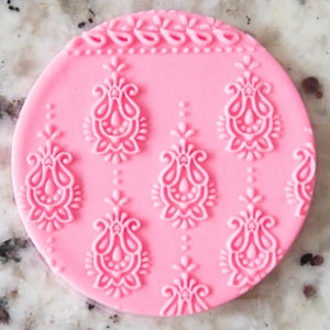 May include: A circular, pink silicone mould with an embossed floral design. The mould features a repeating pattern of stylized teardrop-shaped floral elements and a decorative border along the top edge. The design is raised, creating a textured surface.