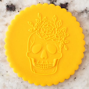 May include: Yellow sugar cookie with a detailed sugar cookie imprint of a skull with flowers on top.