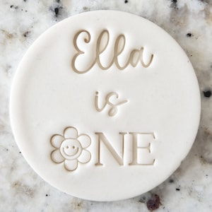 May include: A circular, off-white disc with the words "Ella is ONE" embossed in a light brown colour. A smiling flower graphic is next to the word "ONE".