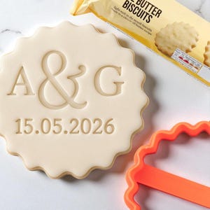 May include: A pale, scalloped-edged biscuit with the initials "A&G" and the date "15.05.2026" imprinted on it. A packet of butter biscuits is in the background, along with an orange cookie cutter.