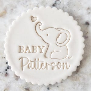 May include: A white, circular baby shower favour with a scalloped edge. The design features a baby elephant with a heart above its trunk and the words "BABY Patterson" engraved in the centre.