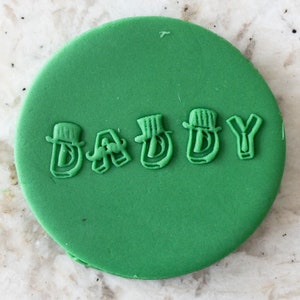 Puede incluir: Galletas de fondant verde con la palabra "DADDY" escrita en glaseado blanco. Cada letra tiene un pequeño sombrero en la parte superior.