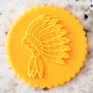 May include: A yellow fondant cookie with a detailed design of a Native American headdress. The headdress is made of feathers and has a beaded band.