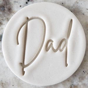 Op de afbeelding: Een ronde, gebroken witte stempel met het woord "Dad" in een cursief lettertype. De letters zijn verzonken in de stempel, waardoor een subtiel, elegant ontwerp ontstaat. De stempel wordt waarschijnlijk gebruikt voor knutselen of bakken.