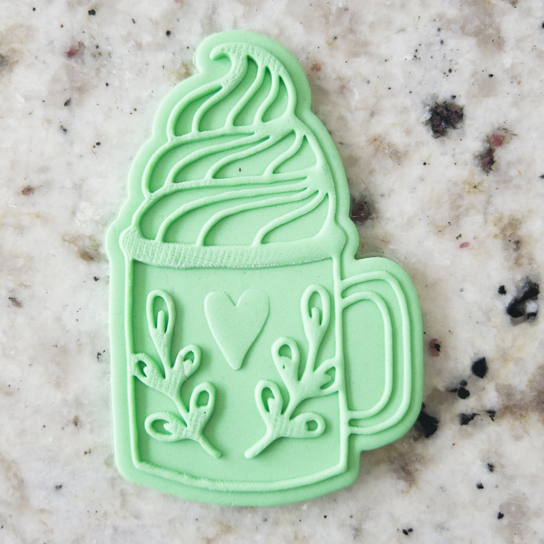 Pode incluir: Um cortador de biscoitos de pl&aacute;stico verde em forma de caneca com chantilly e um desenho de cora&ccedil;&atilde;o. A caneca tem folhas na lateral.