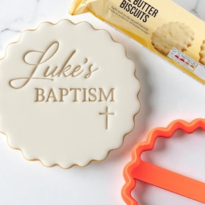 May include: A white, scalloped-edge cookie with the words "Luke's BAPTISM" and a cross design. The cookie has a gold border. A package of butter biscuits and an orange cookie cutter are also in the image.