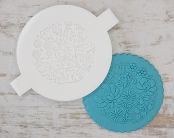 Puede incluir: Una plantilla de decoración de pasteles de plástico blanco con un diseño floral. La plantilla se muestra junto a un molde de decoración de pasteles de silicona azul turquesa con el mismo diseño floral.