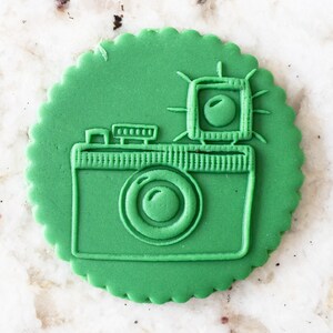 Puede incluir: Una galleta verde decorada con un contorno verde de una cámara con flash. La galleta tiene un borde festoneado.