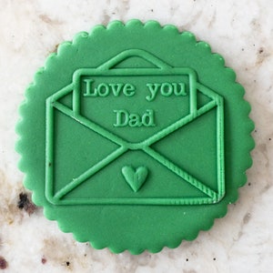 Puede incluir: Galletas verdes con borde festoneado, decoradas con una imagen en relieve de un sobre con un corazón y el texto "Love you Dad".