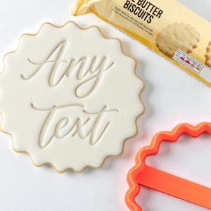 Puede incluir: Una galleta blanca con bordes festoneados con las palabras "Any Text" en escritura cursiva. También se ven un cortador de galletas naranja y un paquete de galletas de mantequilla. La galleta tiene un borde marrón claro.