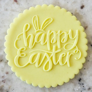 Könnte beinhalten: Ein gelber Fondant-Kuchentopper mit den Worten "Frohe Ostern" in einer kursivschriftlichen Schriftart. Der Topper hat auch ein Paar Hasenohren.