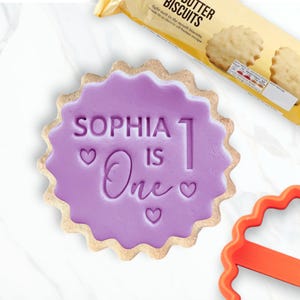 Può includere: Un biscotto decorato con glassa viola con la scritta "SOPHIA 1 IS One" e cuori. Il biscotto ha un bordo smerlato. Sullo sfondo è visibile una confezione di biscotti al burro.