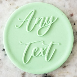 May include: A light green round stamp with the words "Any Text" in a cursive font.