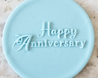 Pode incluir: Um carimbo de fondant circular azul claro com as palavras "Happy Anniversary" em relevo em branco. O texto está em uma fonte cursiva. O carimbo está em uma superfície de mármore branco.