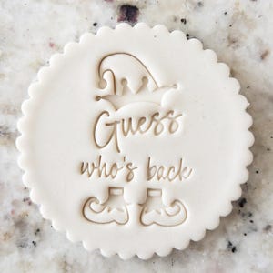 Puede incluir: Cortador de galletas blanco con borde festoneado, que presenta un duende con un sombrero y el texto "Guess who's back".