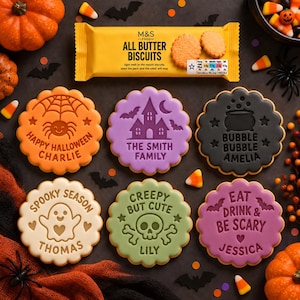 May include: Six round scalloped biscuits with Halloween designs and names, including a spider, haunted house, cauldron, ghost, skull, and bats. The biscuits are arranged below a yellow M&S Food All Butter Biscuits packet, surrounded by pumpkins.