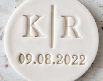 May include: White round ceramic coaster with the initials "K" and "R" separated by a vertical line and the date "09.08.2022" engraved in the center.