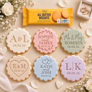 May include: Six round scalloped cookies with embossed text and dates, including A & L, Sophie and James, Mr & Mrs Roberts, E & M, Katie and Josh, and L | K. Above them sits a yellow M&S Food All Butter Biscuits package and a small white ring box.