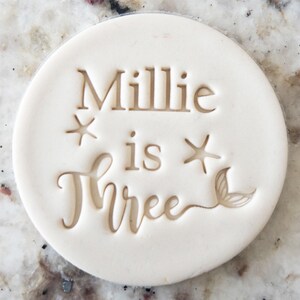 May include: White round cookie cutter with the text "Millie is Three" and a starfish and mermaid tail design.