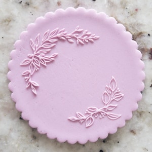 May include: A light pink round cake topper with a white floral design. The design features two sprigs of leaves and berries.