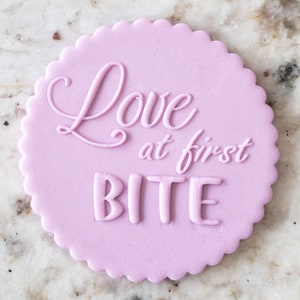Puede incluir: Una galleta redonda de color morado claro con el texto "Love at first bite" en glaseado blanco.