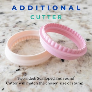 May include: Two pink plastic cookie cutters, one with a scalloped edge and one with a smooth round edge. The text "ADDITIONAL CUTTER" is above the cutters. The text "Two sided: Scalloped and round Cutter will match the chosen size of stamp." is below the cutters.