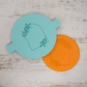 May include: Two stencils for decorating cakes or cookies. One stencil is light blue and the other is orange. Both stencils feature a rectangular frame with leafy branches on either side.
