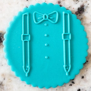 Puede incluir: Un cortador de fondant o galletas de color turquesa con forma de esmoquin. El diseño incluye una pajarita, tirantes y botones, todo delineado en un círculo festoneado.