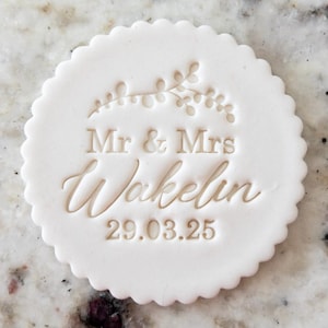 Może przedstawiać: Biały, okrągły topper na tort z falistym brzegiem. Topper jest wytłoczony wzorem kwiatowym, tekstem "Mr & Mrs Wakelin" i datą "29.03.25".