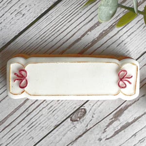 Puede incluir: Una galleta rectangular blanca con un borde dorado y dos lazos rojos en cada extremo. La galleta está en blanco y lista para ser decorada.