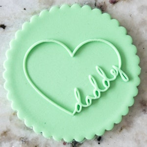 Puede incluir: Una galleta redonda de color verde claro con un corazón blanco y la palabra "papá" escrita dentro del corazón.