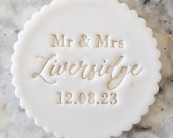 May include: White round cake topper with the text "Mr & Mrs Liveridge 12.08.23" embossed in gold.
