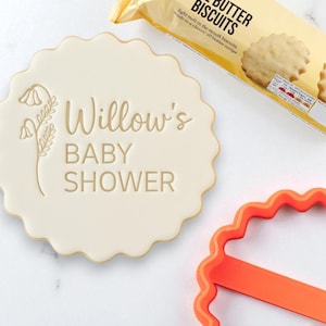 Può includere: Un biscotto beige con bordo smerlato con la scritta "Willow's BABY SHOWER" e un motivo floreale. Una confezione di biscotti al burro è sullo sfondo, insieme a un tagliabiscotti arancione.