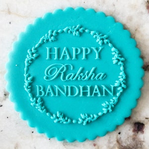 Puede incluir: Una galleta redonda de color azul turquesa con un borde festoneado y una inscripción blanca que dice "Happy Raksha Bandhan" rodeada de una corona de diseños florales blancos.