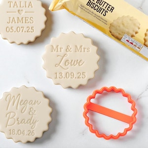May include: Close-up of personalized cookies with names and dates, including "Talia James 05.07.25", "Mr & Mrs Lowe 13.09.25", and "Megan & Brady 18.04.26". An orange cookie cutter and a package of butter biscuits are also visible.