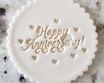 Pode incluir: Topo de bolo redondo branco com as palavras "Happy Anniversary!" em letras douradas e pequenos recortes em forma de coração ao redor do texto.