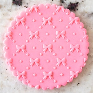May include: A pink fondant round cake topper with a repeating pattern of bows and dots.