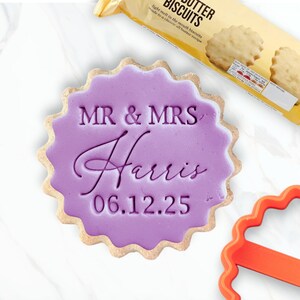 May include: A wedding-themed biscuit with a purple icing centre, inscribed with "MR & MRS Harris 06.12.25". The biscuit has a scalloped edge. A packet of butter biscuits and an orange cookie cutter are also visible.