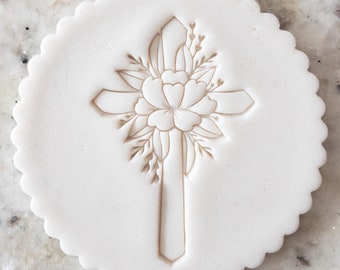 May include: A white, round, scalloped-edge cookie cutter with a floral design featuring a cross and a large flower.