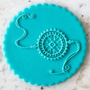 Puede incluir: Un pastel redondo azul turquesa decorado con un diseño blanco de encaje de un brazalete Rakhi tradicional indio. El brazalete está atado con un nudo y tiene un amuleto circular en el centro.